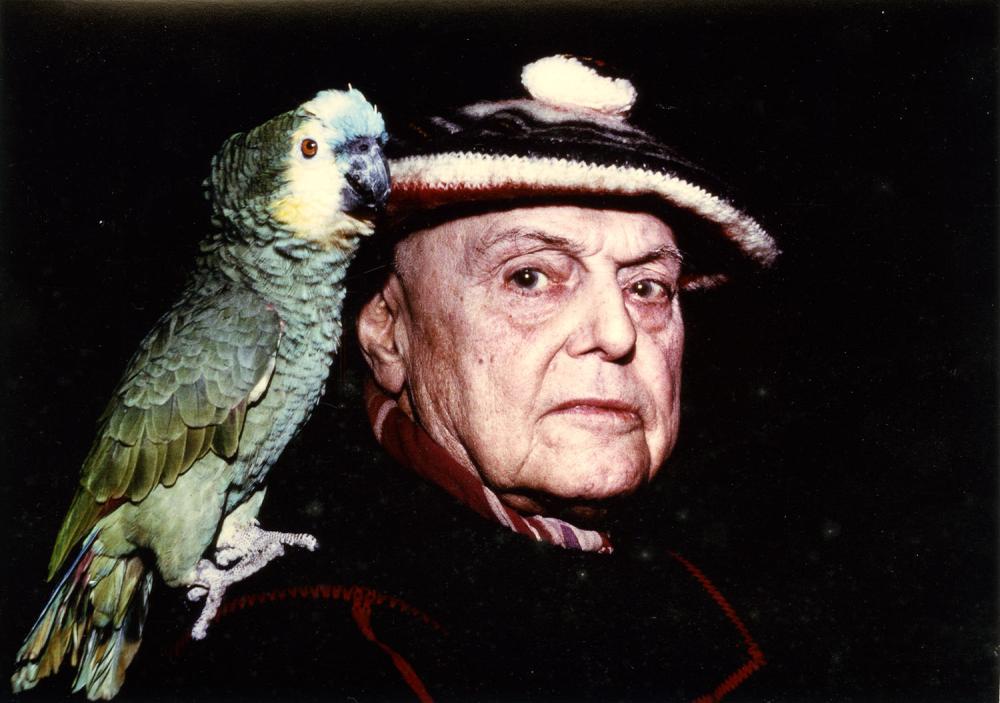 Photographie couleur représentant, sur un fond noir, un gros plan de l'écrivain Pierre Mac Orlan avec son perroquet posé sur l'épaule. Le perroquet est vert, Mac Orlan porte un béret.