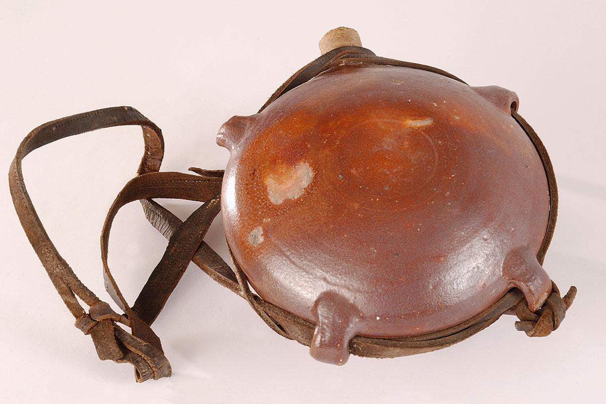 Gourde en grès, de forme sphérique, aux parois un peu aplaties, comportant une rainure sur le pourtour et des "passants" en grès pour permettre le passage d'un lien en cuir. La gourde est fermée par un bouchon de liège.