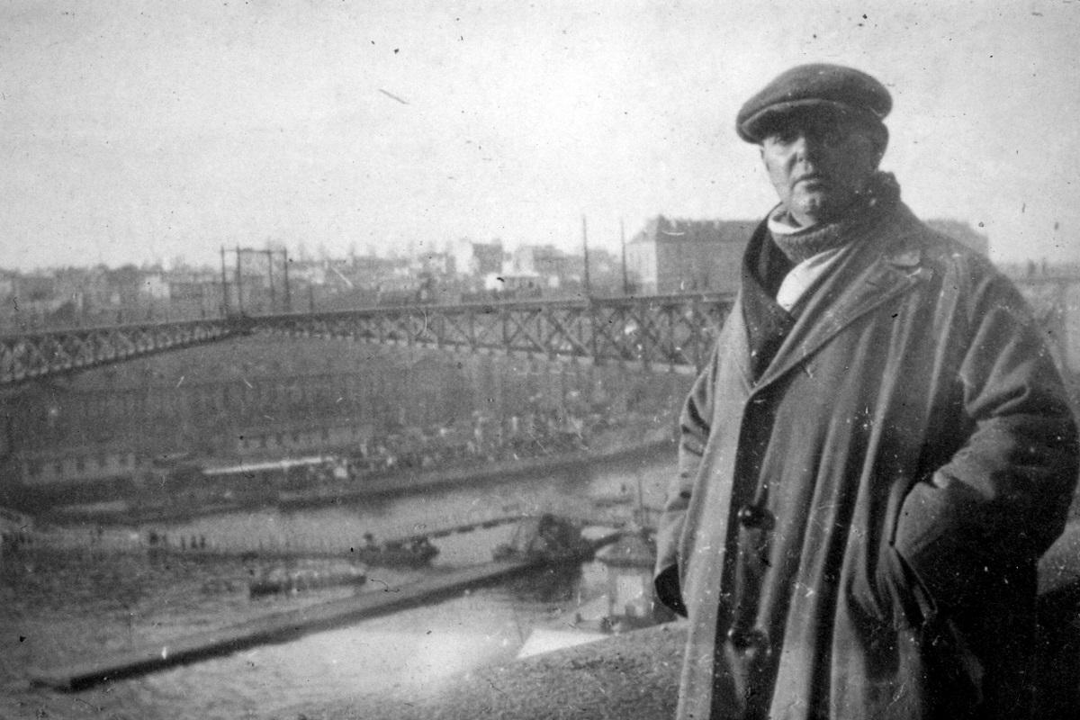 Photographie monochrome, ancienne, la surface est usée et les contrastes estompés. Pierre Mac Orlan porte un imperméable et une casquette, il surplombe un port, à l'arrière un pont métallique enjambe la zone d'activité.