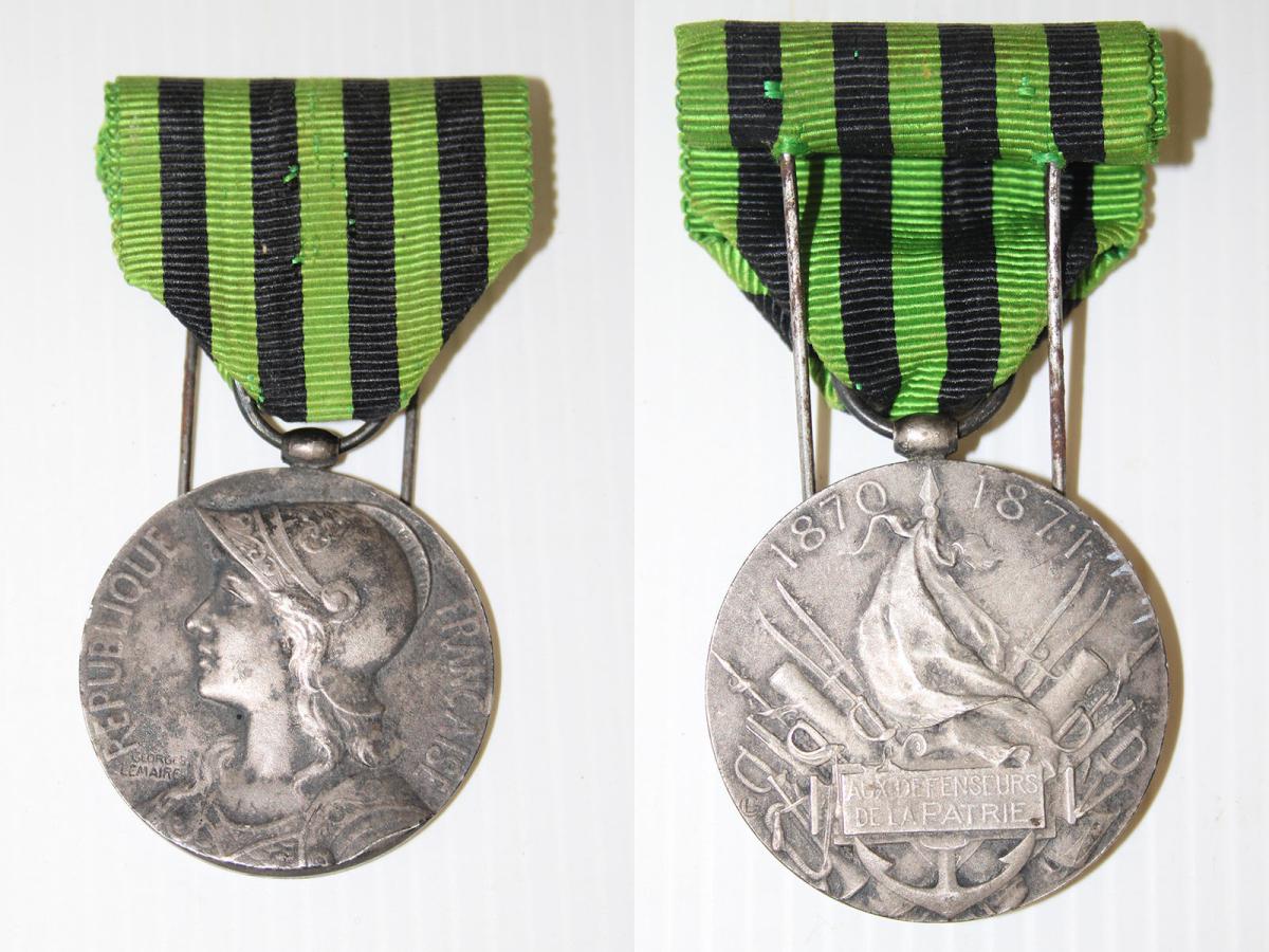 Médaille en métal, 1 face représente Marianne casquée, l'autre face porte l'inscription "Aux défenseurs de la Patrie" et le millésime 1870-1871 ; ruban en tissu rayé vert et noir