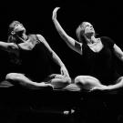 Photographie en noir et blanc. Deux danseuses, assises côte à côte, exécutent le même mouvement : le bras droit relevé, arrondi, le buste incliné de leur mouvement.
