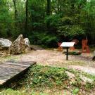 Photographie en couleur réalisée au Bois de la Barre à la Ferté-sous-Jouarre : un panneau explicatif,une meule, des pierres meulières et des silhouettes de carriers agrémentent la promenade.