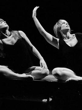 Photographie en noir et blanc. Deux danseuses, assises côte à côte, exécutent le même mouvement : le bras droit relevé, arrondi, le buste incliné de leur mouvement.