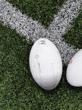Deux ballons de rugby posés sur une ligne blanche tracée sur un terrain herbeux. Dans le coin en bas à gauche, le bout de deux chaussures de sport blanchesgauchesur 