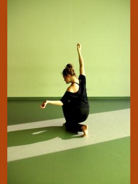 Photographie de Camille Desmarest en train de danser, de dos, le genou posé au sol, la jambe droite pliée devant elle, le bras droit levé, tendu et le bras gauche est replié, l'avant bras ouvert vers l'extérieur. Elle porte un ensemble noir, ses cheveux sont relevés en chignon. L'éclairage vert