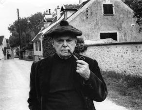 Photographie monochrome de Pierre Mac Orlan. Portrait en buste de l'écrivain, fumant la pipe, sur la route au hameau d'Archets, avec à l'arrière-plan sa maison