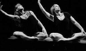 Photographie en noir et blanc. Deux danseuses, assises côte à côte, exécutent le même mouvement : le bras droit relevé, arrondi, le buste incliné de leur mouvement.
