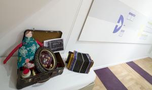 Photographie couleur de l'entrée de l'exposition, au premier plan une valise ouverte contenant une poupée en tissu, un cadre avec des médailles, une photo en noir et blanc, un pot en terre cuite, posé à côté un petit accordéon.