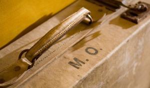 Gros plan de la poignée et des initiales M.O. sur une valise adossée à un mur jaune