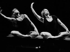 Photographie en noir et blanc. Deux danseuses, assises côte à côte, exécutent le même mouvement : le bras droit relevé, arrondi, le buste incliné de leur mouvement.