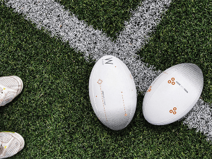 Deux ballons de rugby posés sur une ligne blanche tracée sur un terrain herbeux. Dans le coin en bas à gauche, le bout de deux chaussures de sport blanchesgauchesur
