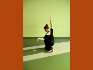 Photographie de Camille Desmarest en train de danser, de dos, le genou posé au sol, la jambe droite pliée devant elle, le bras droit levé, tendu et le bras gauche est replié, l'avant bras ouvert vers l'extérieur. Elle porte un ensemble noir, ses cheveux sont relevés en chignon. L'éclairage vert