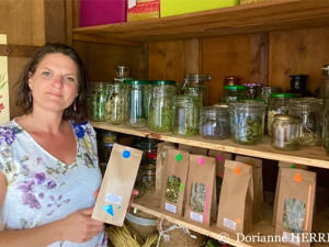 Photographie couleur de l'herboriste Dorianne Herrera, debout devant une étagère sur laquelle sont présentés les produits qu'elle propose à la vente, dans des bocaux en verre et des sachets en papiers