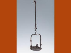 Lampe à huile en métal, de forme circulaire avec une ouverture condamnée par une une plaque ornée d'un petit coq. La lampe est munie d'une tige d'anse mobile et de suspension à crochet.