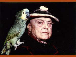 Photographie couleur représentant, sur un fond noir, un gros plan de l'écrivain Pierre Mac Orlan avec son perroquet posé sur l'épaule. Le perroquet est vert, Mac Orlan porte un béret.