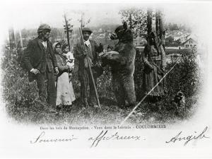 Carte postale représentant un groupe de personne dans des bois, entourant un ours debout sur les pattes arrières et portant une muselière ; la femme sur la droite tient un petit singe en laisse