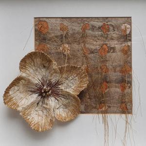 Photographie d'une création de Lola Greenwich : une grande fleur en fibres naturelles, à cinq pétales de couleur crème, rebrodées sur les bords extérieurs de fil marron clair, le pistil est en perles, au centre d'une broderie marron. La fleur est posée sur le bord d'un cadre carré également en fibres, dans les tons beige, marron et orange.
