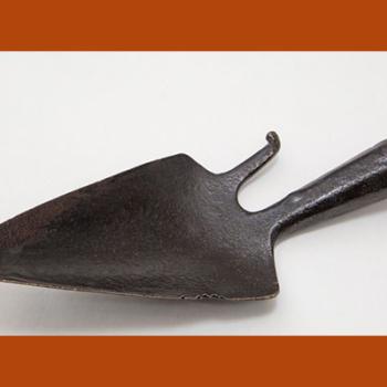 Outil en fer forgé, en forme de petite pelle pointue avec un crochet à droite. Le manche est manquant.