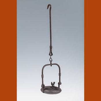 Lampe à huile en métal, de forme circulaire avec une ouverture condamnée par une une plaque ornée d'un petit coq. La lampe est munie d'une tige d'anse mobile et de suspension à crochet.
