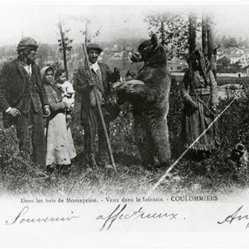 Carte postale représentant un groupe de personne dans des bois, entourant un ours debout sur les pattes arrières et portant une muselière ; la femme sur la droite tient un petit singe en laisse
