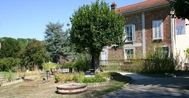 Meule en pierre, carrés de plantes utiles, tilleuls et barrière en osier devant l'entrée de la cour du Musée de la Seine-et-Marne.