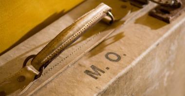 Gros plan de la poignée et des initiales M.O. sur une valise adossée à un mur jaune