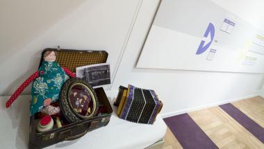 Photographie couleur de l'entrée de l'exposition, au premier plan une valise ouverte contenant une poupée en tissu, un cadre avec des médailles, une photo en noir et blanc, un pot en terre cuite, posé à côté un petit accordéon. 