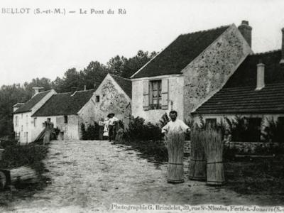Carte postale ancienne du village de Bellot en Seine-et-Marne, au 1er plan un vannier avec des bottes d'osier posées devant lui.