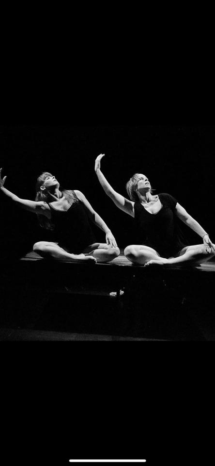 Photographie en noir et blanc. Deux danseuses, assises côte à côte, exécutent le même mouvement : le bras droit relevé, arrondi, le buste incliné de leur mouvement.