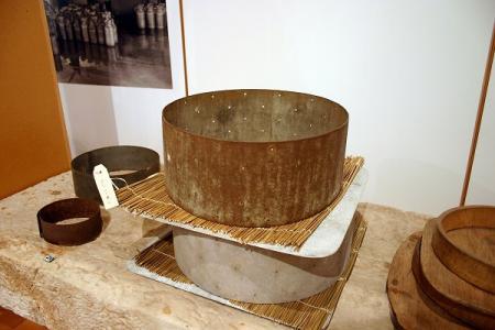 vue partielle de la table d’égouttage (n° inventaire1983.8.1), avec des moules à fromage de différents diamètres et matériaux (métal, bois) posés dessus.