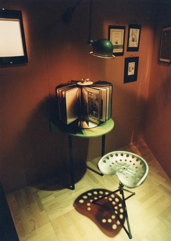 Dans l'espace "Usines et Machines", un présentoir vertical contenant de nombreuses photos est posé sur une console devant un tabouret d'atelier en métal.