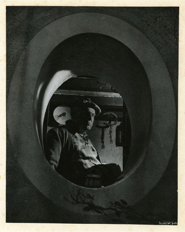 Portrait en noir et blanc de Pierre Mac Orlan par Michel Cot. Il se tient dans l'obscurité devant la fenêtre dite "œil de bœuf" dans sa maison