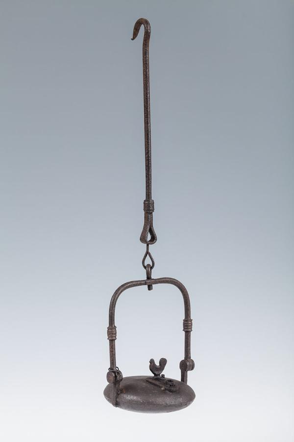 Lampe à huile en métal, de forme circulaire avec une ouverture condamnée par une une plaque ornée d'un petit coq. La lampe est munie d'une tige d'anse mobile et de suspension à crochet.
