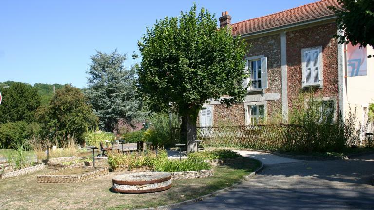 Meule en pierre, carrés de plantes utiles, tilleuls et barrière en osier devant l'entrée de la cour du Musée de la Seine-et-Marne.