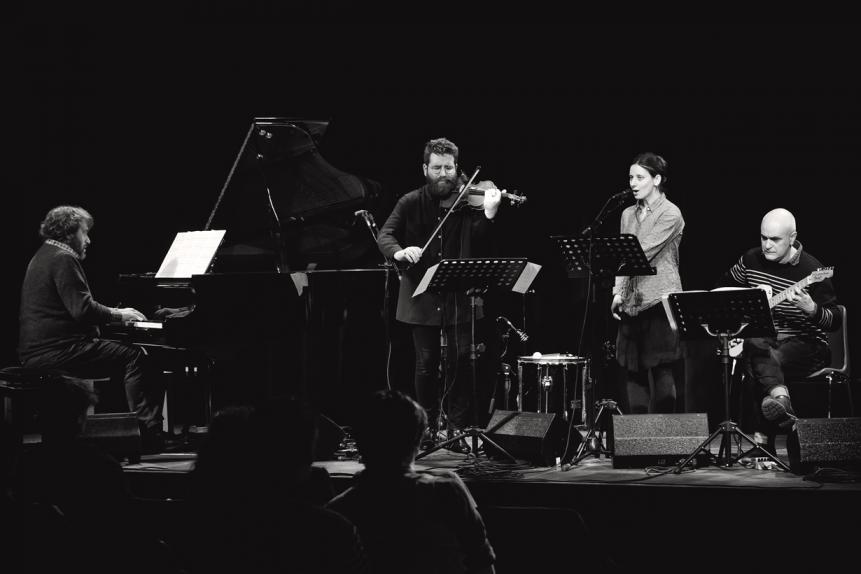 Photographie en noir et blanc de l'ensemble Dedalus en concert. Le groupe est composé de trois musiciens qui jouent du piano, du violon et de la guitare et d'une chanteuse