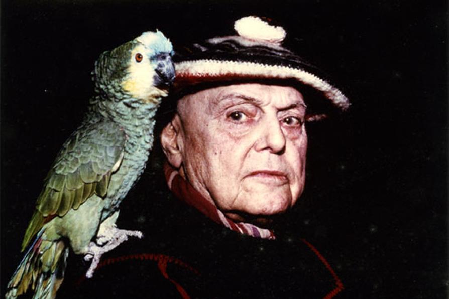 Photographie couleur représentant, sur un fond noir, un gros plan de l'écrivain Pierre Mac Orlan avec son perroquet posé sur l'épaule. Le perroquet est vert, Mac Orlan porte un béret.