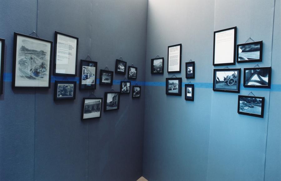 Sur un mur bleu des photos monochromes de voitures, notamment de Pierre Mac Orlan avec différentes automobiles, surle côté droit, une voiture de course ancienne. en 