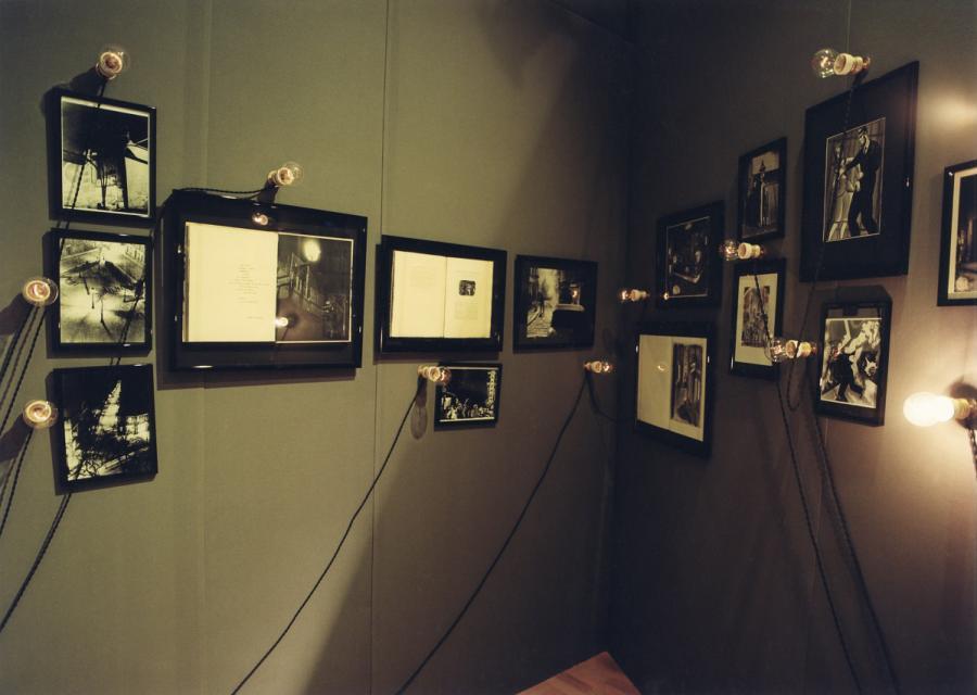Espace "Électicité", sur un mur vert kaki, des photos monochromes et des livres encadrés, accrochés au mur et surmontés ou entourés d'ampoules électriques. Les fils électriques ont été délibérément laissés apparents 
